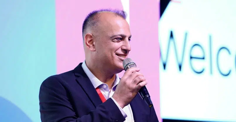 גבר בחליפה מדבר למיקרופון על הבמה, עומד מול רקע צבעוני שבו ניתן לראות חלק מהמילה "Welcome" (ברוכים הבאים).